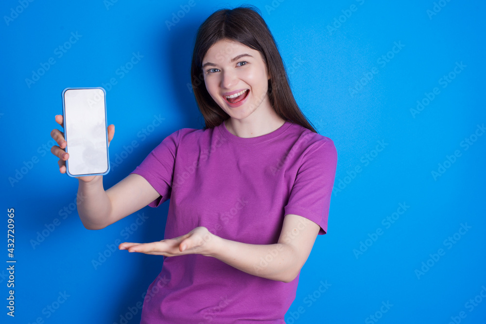 young beautiful Caucasian woman wearing purple T-shirt over blue wall with a mobile. presenting smartphone. Advertisement concept. young beautiful Caucasian woman wearing purple T-shirt over blue wall with a mobile. presenting smartphone. Advertisement concept.