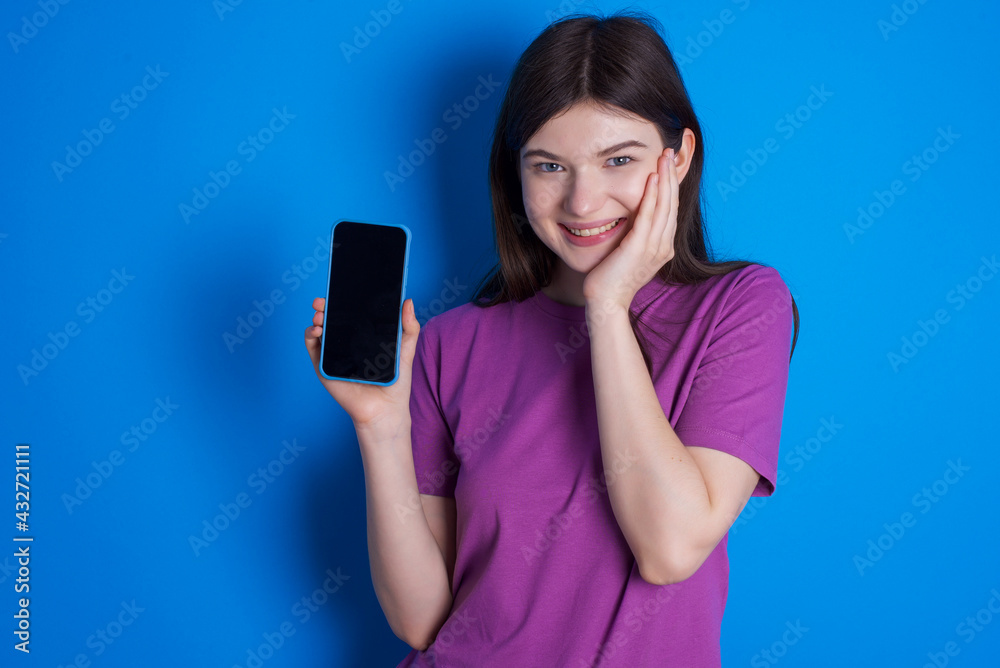young beautiful Caucasian woman wearing purple T-shirt over blue wall hold hand modern technology use touch face palm astonished impressed scream wow omg unbelievable unexpected young beautiful Caucasian woman wearing purple T-shirt over blue wall hold hand modern technology use touch face palm astonished impressed scream wow omg unbelievable unexpected