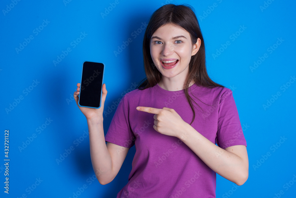 Attractive cheerful young beautiful Caucasian woman wearing purple T-shirt over blue wall holding in hands cell showing black screen Attractive cheerful young beautiful Caucasian woman wearing purple T-shirt over blue wall holding in hands cell showing black screen