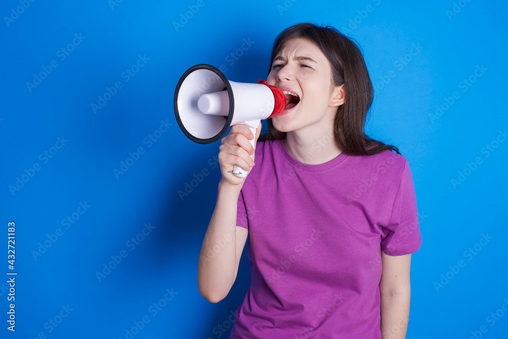 young beautiful Caucasian woman wearing purple T-shirt over blue wall Through Megaphone with Available Copy Space young beautiful Caucasian woman wearing purple T-shirt over blue wall Through Megaphone with Available Copy Space