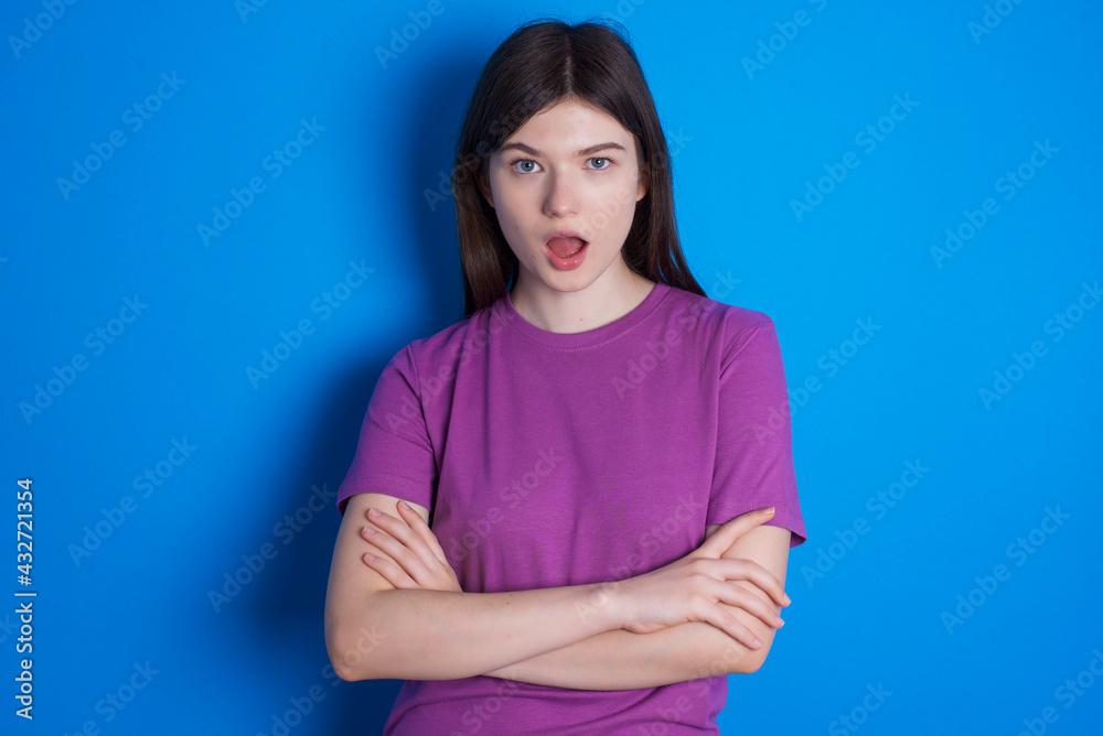 Shocked embarrassed young beautiful Caucasian woman wearing purple T-shirt over blue wall keeps mouth widely opened. Hears unbelievable novelty stares in stupor Shocked embarrassed young beautiful Caucasian woman wearing purple T-shirt over blue wall keeps mouth widely opened. Hears unbelievable novelty stares in stupor
