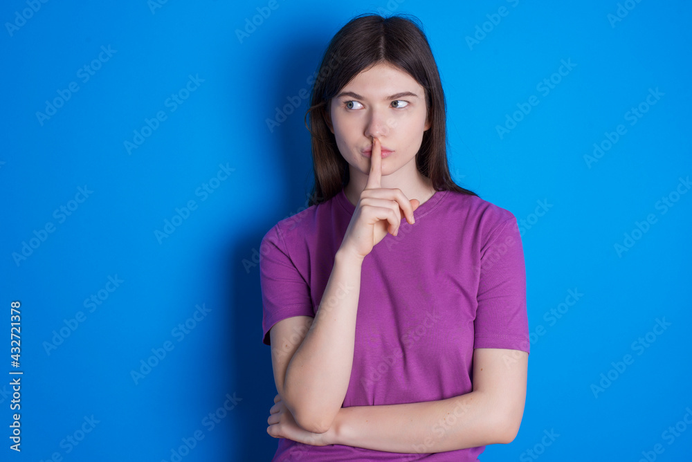 young beautiful Caucasian woman wearing purple T-shirt over blue wall silence gesture keeps index finger to lips makes hush sign. Asks not to share secret young beautiful Caucasian woman wearing purple T-shirt over blue wall silence gesture keeps index finger to lips makes hush sign. Asks not to share secret