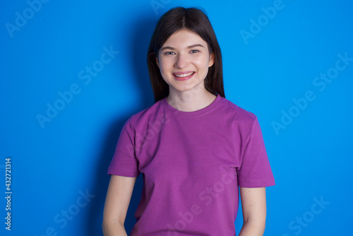 young beautiful Caucasian woman wearing purple T-shirt over blue wall with nice beaming smile pleased expression. Positive emotions concept © Roquillo