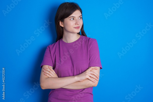 Pleased young beautiful Caucasian woman wearing purple T-shirt over blue wall keeps hands crossed over chest looks happily aside