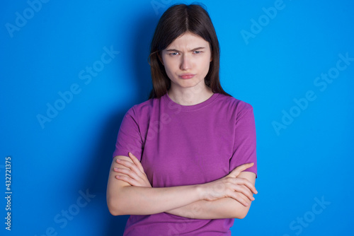 Gloomy dissatisfied young beautiful Caucasian woman wearing purple T-shirt over blue wall looks with miserable expression at camera from under forehead, makes unhappy grimace