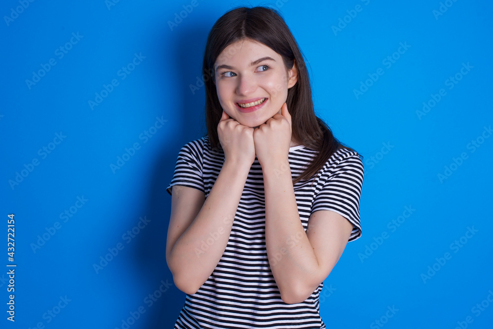 young beautiful Caucasian woman wearing pink jacket over blue wall holds hands under chin, glad to hear heartwarming words from stranger young beautiful Caucasian woman wearing pink jacket over blue wall holds hands under chin, glad to hear heartwarming words from stranger