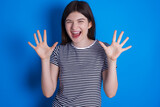 Delighted positive young beautiful Caucasian woman wearing stripped T-shirt over blue wall opens mouth  and arms palms up after having great result