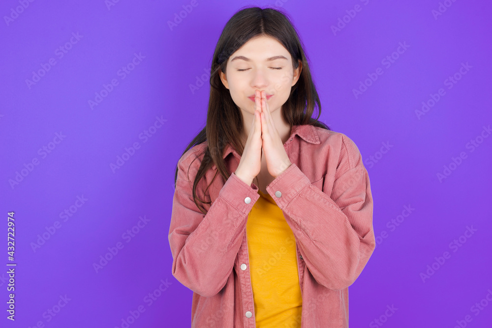Indoor closeup of young beautiful Caucasian woman wearing casual clothes over purple wall practicing yoga and meditation, holding palms together in namaste, looking calm, relaxed and peaceful. Indoor closeup of young beautiful Caucasian woman wearing casual clothes over purple wall practicing yoga and meditation, holding palms together in namaste, looking calm, relaxed and peaceful.