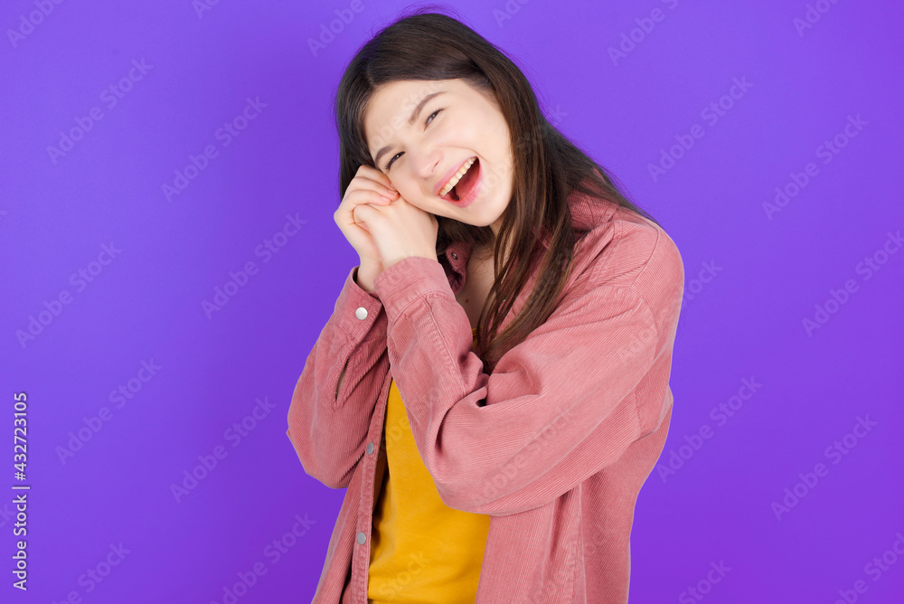 Dreamy young beautiful Caucasian woman wearing casual clothes over purple wall with pleasant expression, closes eyes, keeps hands crossed near face, thinks about something pleasant Dreamy young beautiful Caucasian woman wearing casual clothes over purple wall with pleasant expression, closes eyes, keeps hands crossed near face, thinks about something pleasant
