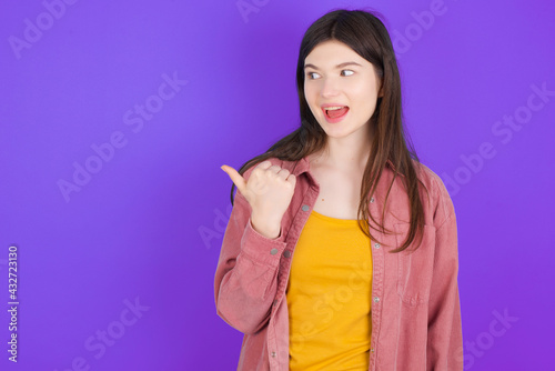 Stupefied young beautiful Caucasian woman wearing casual clothes with surprised expression, opens eyes and mouth widely, points aside with thumb, shows something strange. Advertisement concept.