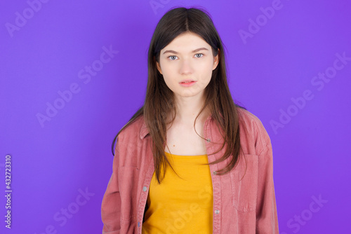Joyful young beautiful Caucasian woman wearing casual clothes over purple wall looking to the camera, thinking about something. Both arms down, neutral facial expression.