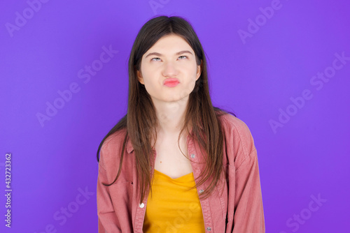 young beautiful Caucasian woman wearing casual clothes over purple wall has worried face looking up lips together, being upset thinking about something important, keeps hands down.