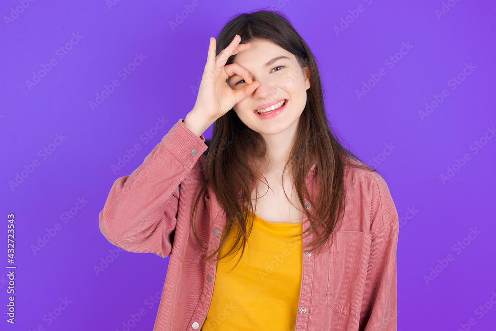 young beautiful Caucasian woman wearing casual clothes over purple wall with happy face smiling doing ok sign with hand on eye looking through finger. young beautiful Caucasian woman wearing casual clothes over purple wall with happy face smiling doing ok sign with hand on eye looking through finger.