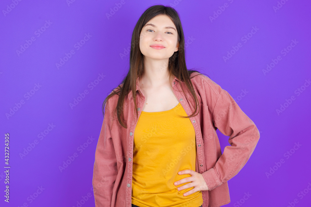 Studio shot of cheerful young beautiful Caucasian woman wearing casual clothes over purple wall keeps hand on hip, smiles broadly. Studio shot of cheerful young beautiful Caucasian woman wearing casual clothes over purple wall keeps hand on hip, smiles broadly.
