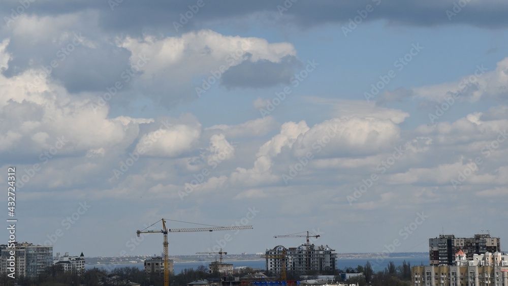 Construction crane jibs over rooftops under cloudscape. Under construction condo buildings in European city. Heavy construction machinery at urban skyline with cumulus clouds Construction crane jibs over rooftops under cloudscape. Under construction condo buildings in European city. Heavy construction machinery at urban skyline with cumulus clouds