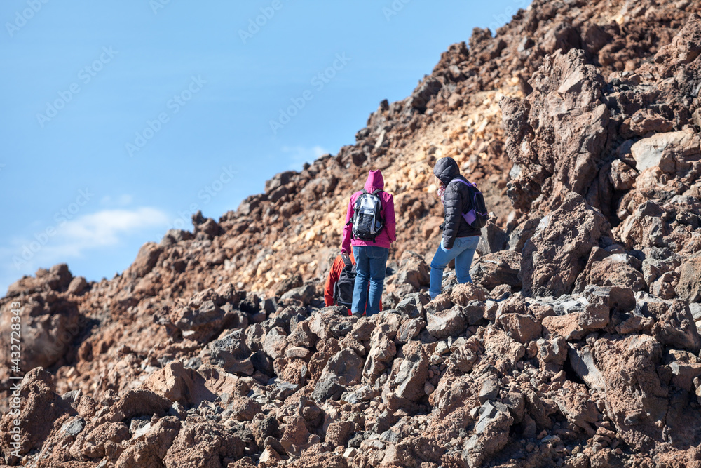 Three hikers descend along a stone path among the lava piles high in the mountains in sunny and clear weather. The Teide Volcano, Tenerife, Canary Islands, Spain