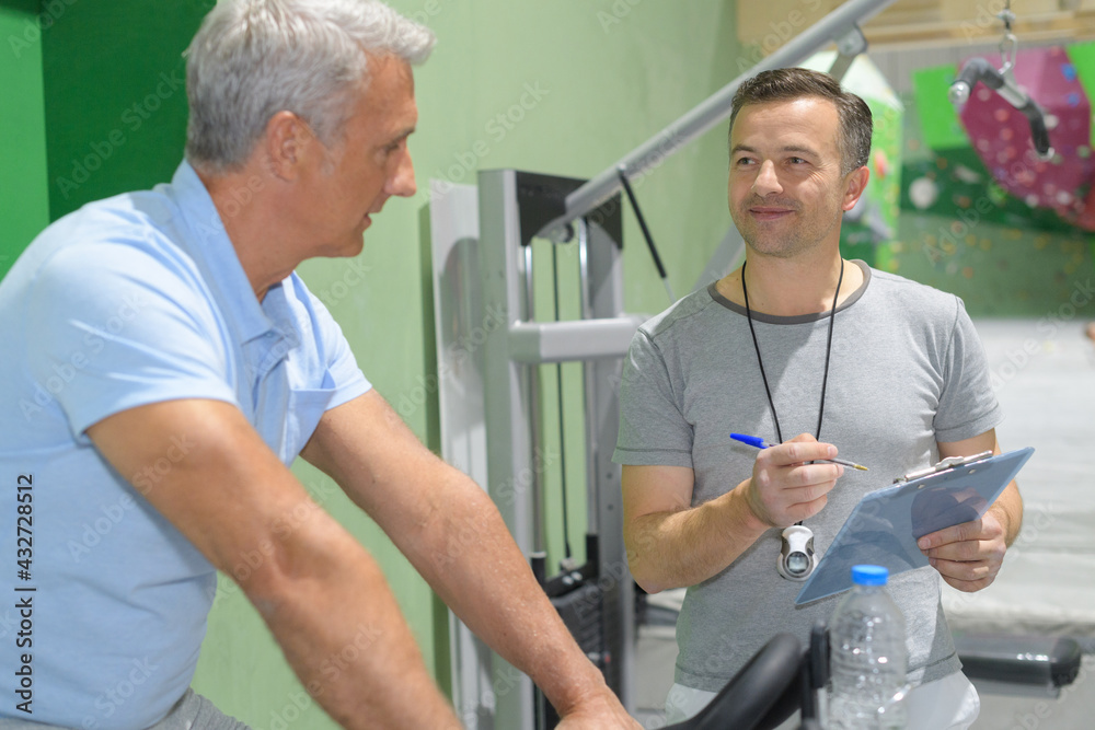 Fototapeta premium personal trainer holding clipboard with senior man using exercise equipment