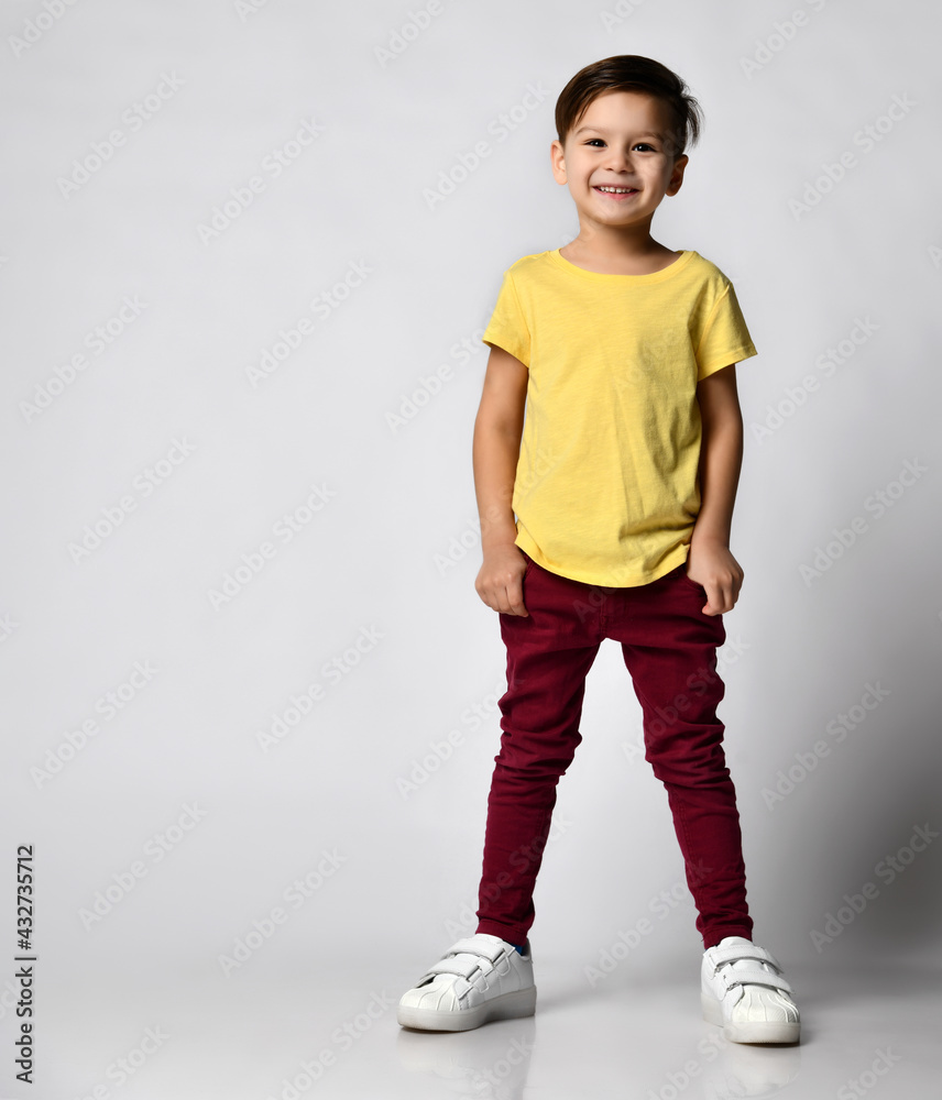 Foto Stock Full body studio portrait of fashionable preschool little boy  wearing yellow t-shirt, red denim pants, white sneakers, standing with  hands in pockets, smiling. isolated on light background | Adobe Stock