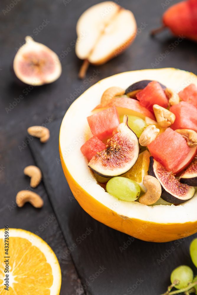 Vegetarian fruit salad of watermelon, grapes, figs, pear, orange, cashew  on black concrete background. Side view, selective focus.