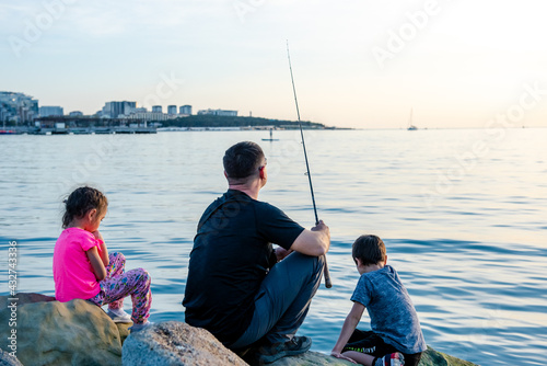 Father with children on a fishing trip by the sea. A boy and a girl with their father have fun fishing on the beach or by the sea. Children fish over a cliff in the sea. Summer sports. 