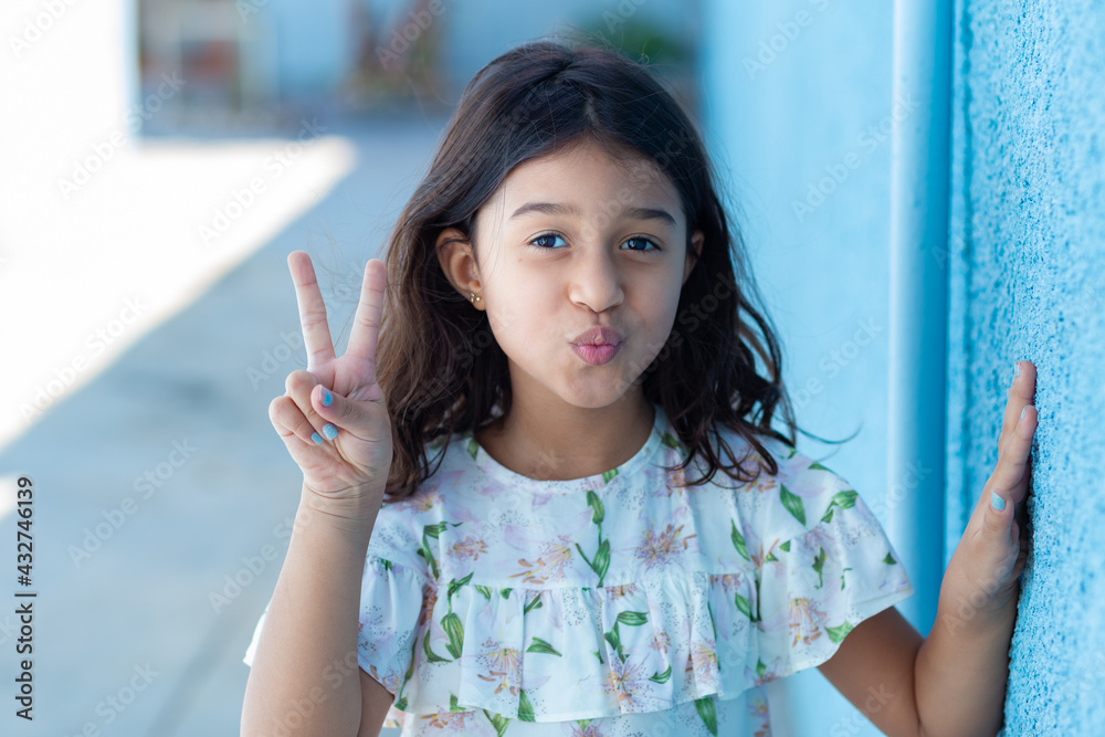 Uma criança, menina, brasileira dos cabelos pretos e lisos, fazendo biquinho encostada no muro pintado de azul. Fazendo sinal de Uma criança, menina, brasileira dos cabelos pretos e lisos, fazendo biquinho encostada no muro pintado de azul. Fazendo sinal de