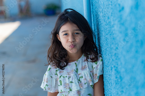 Uma criança, menina, brasileira dos cabelos pretos e lisos, fazendo biquinho encostada no muro pintado de azul. photo