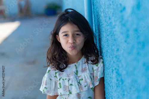 Uma criança, menina, brasileira dos cabelos pretos e lisos, fazendo biquinho encostada no muro pintado de azul. photo