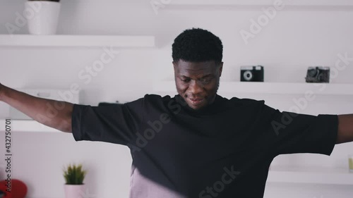 Footage of a guy smiling and wearing a black shirt warming up for a workout. Moving arms in a circular motion. Stretching. White shelves in the background with red boxing gloves and a pot plant.  photo