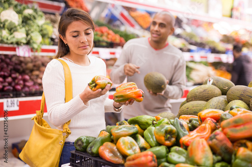 Young latin american girl choosing sweet pepper in supermarket with her boyfriend