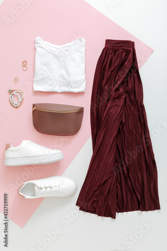 Female spring look summer outfit burgundy skirt white shoes sneakers white basic tshirt waist bag. Folded fly clothes for women fashion urban basic outfit with accessories on pink background. Top view