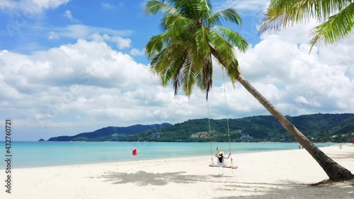 4K Phuket, Thailand. Tropical beach paradise with beach swing with girl in white shirt. Women relax on swing under coconut palm tree at beautiful tropical beach White sand.