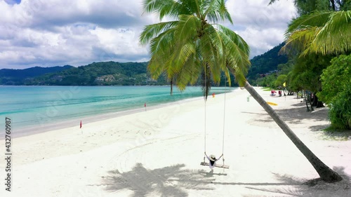 4K Phuket, Thailand. Tropical beach paradise with beach swing with girl in white shirt. Women relax on swing under coconut palm tree at beautiful tropical beach White sand.