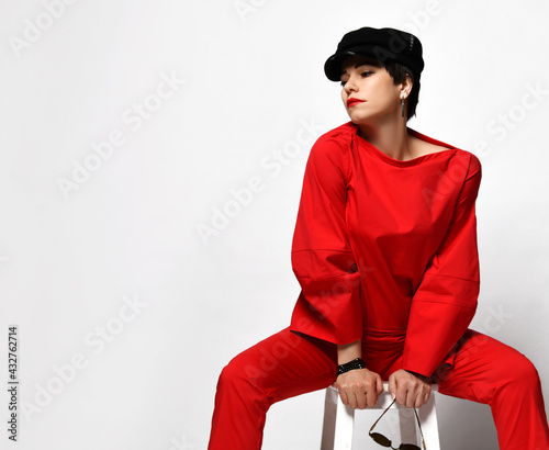Принти на полотні Adult pretty sensual short haired brunette woman in stylish casual red pantsuit and black cap sits on chair with her head turned aside over grey background