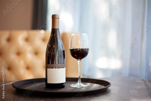 A bottle of red wine and a full glass on a tray stand on a table in the restaurant. Empty label template.