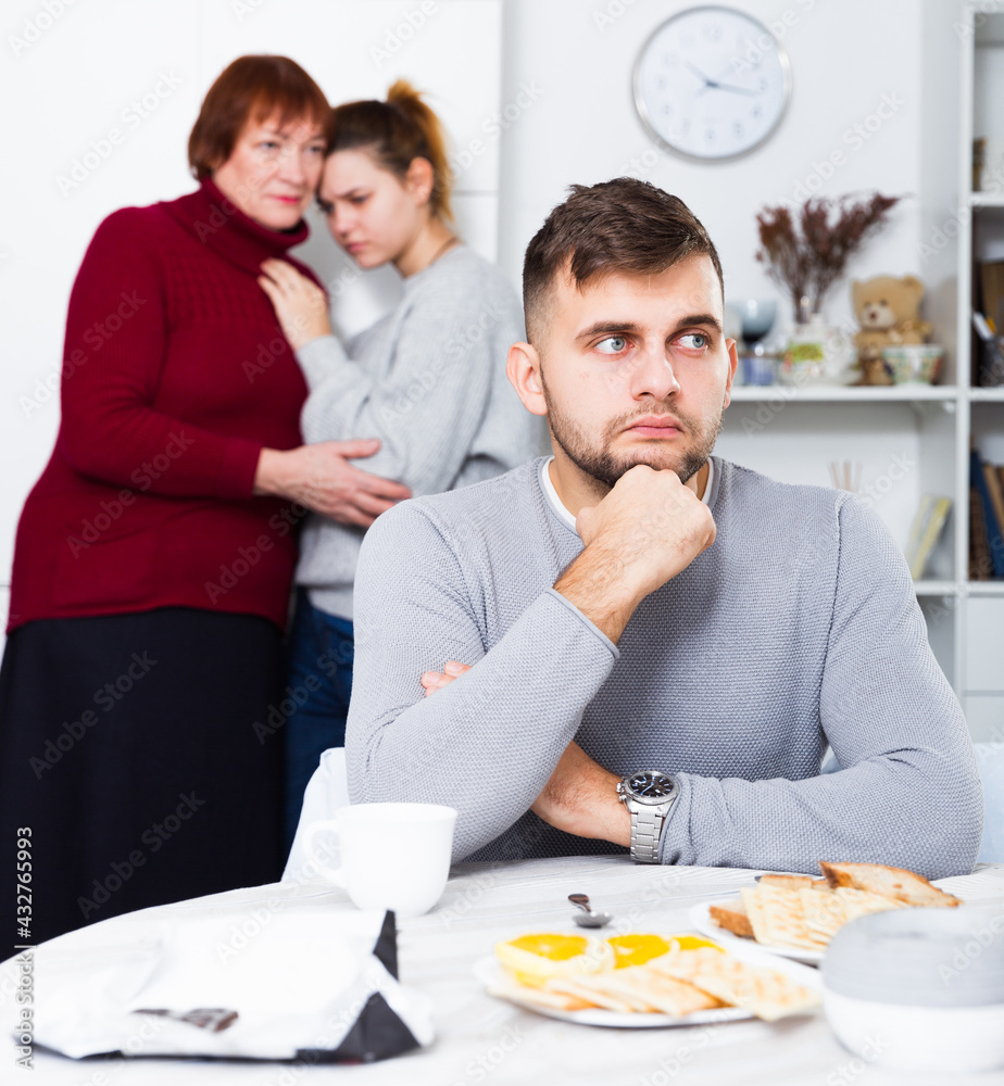 Young upset man sitting separately having problems in relationship with wife and her mother