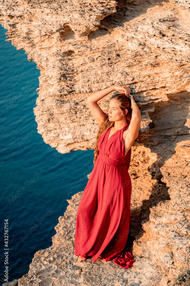 A girl with loose hair in a red dress stands on a rock rock above the sea. In the background, the sea and the rocks. The concept of travel
