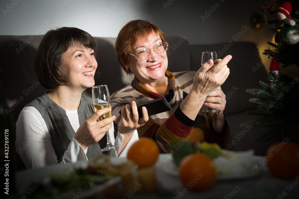 Smiling women watching Christmas tv program sitting at table with wine glasses in dark room