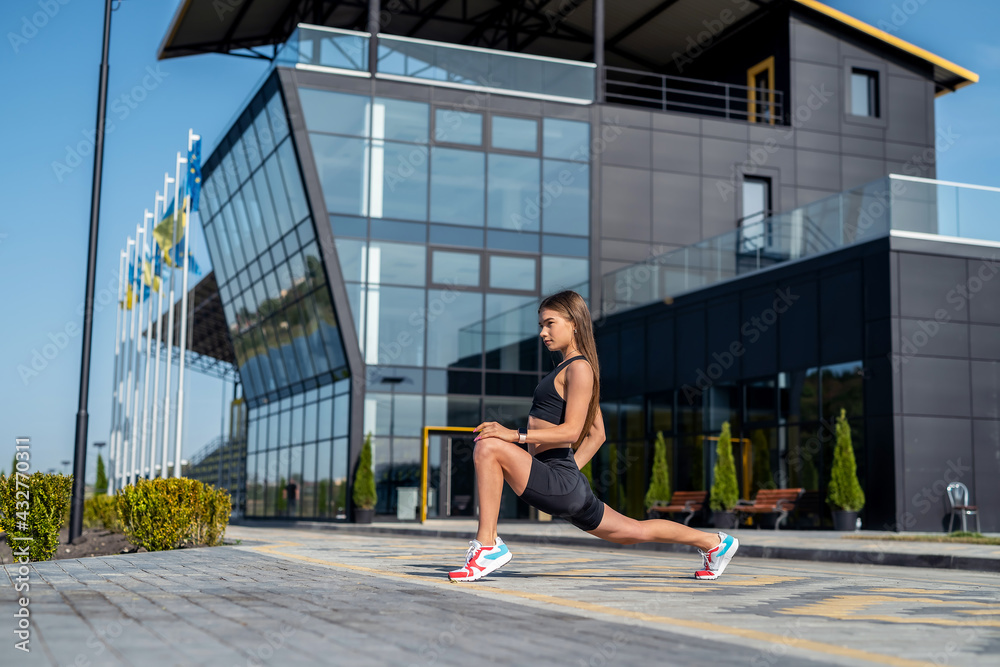 Beautiful young slim woman in sporwear doing morning exercises near modern glass building