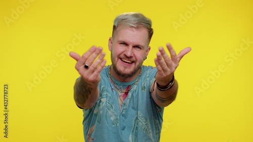 Come to me, I want to embrace you. Stylish tourist guy in t-shirt spread hands and give hug to you. Pleasant expression, love feelings. Young man on yellow studio background. People sincere emotions