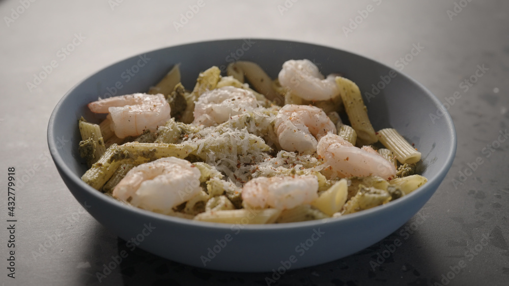 grate cheese on pesto penno with shrimps in blue bowl on concrete countertop