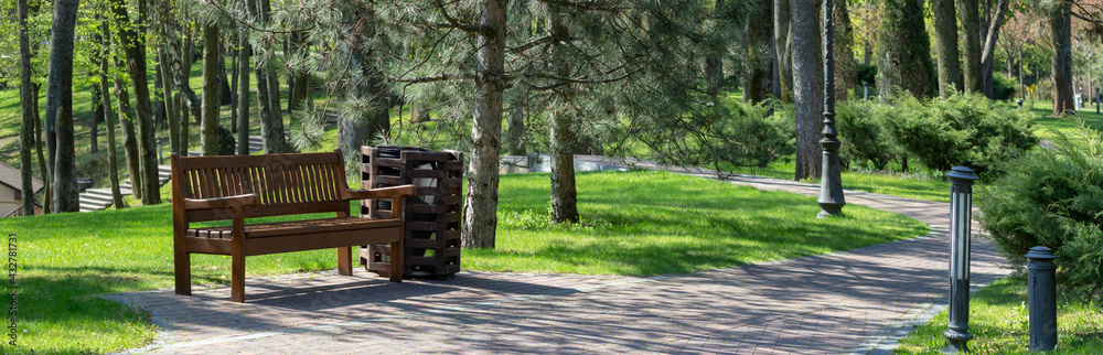 Park bench under a tree in the shade. Trash can near the bench. Path for a walk in the sunny park in the spring. Park bench under a tree in the shade. Trash can near the bench. Path for a walk in the sunny park in the spring.