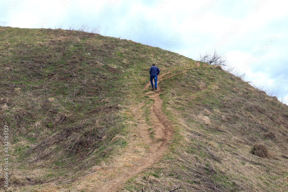 A man in a blue jacket and jeans climbs up a steep mountain-the concept of spring walks in a picturesque area