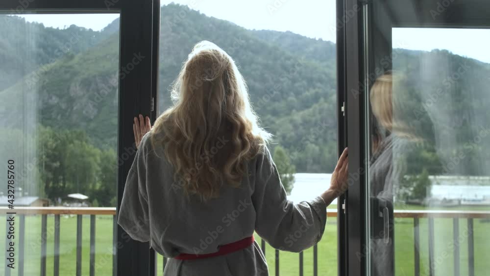 a woman in a bathrobe stands with her back at the window overlooking the mountains