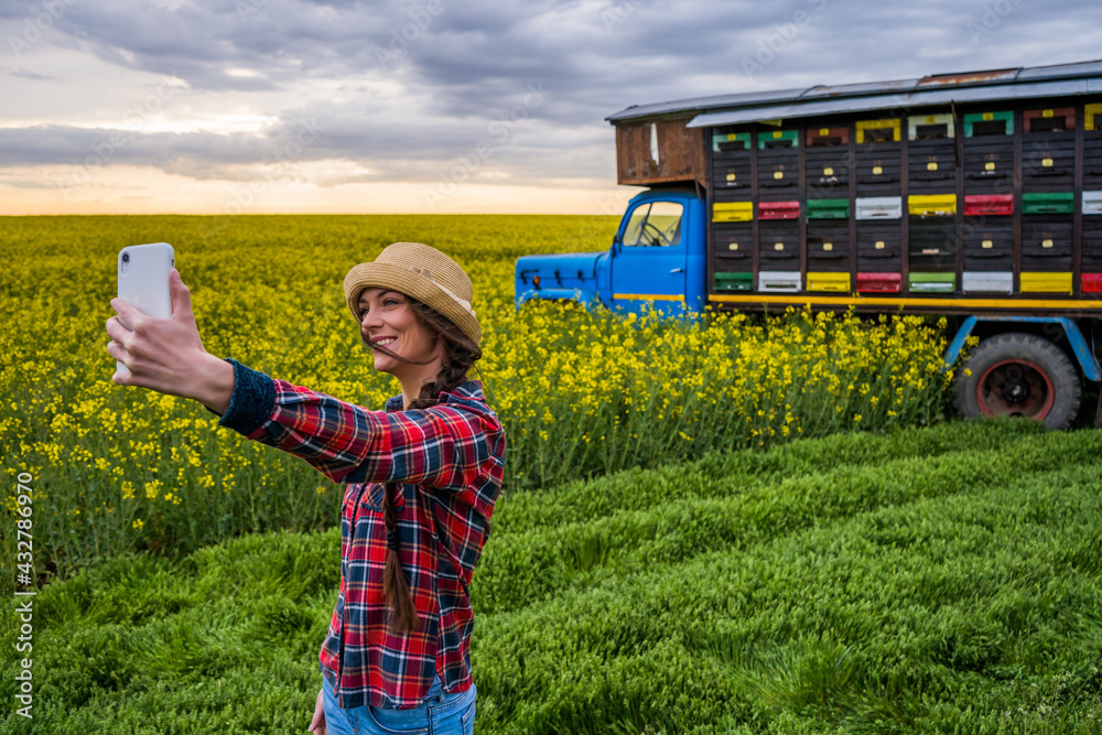 Obraz premium Proud female beekeeper is taking selfie in front of her truck with beehives.