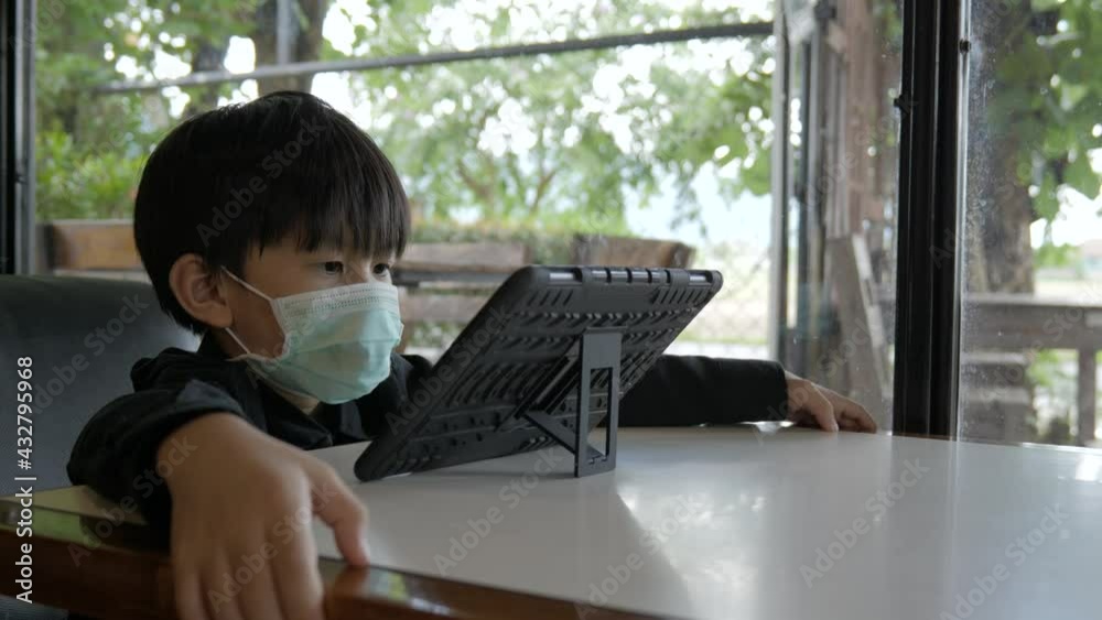 Asian boy watching computer tablet with a medical face mask to prevent the covid-19 virus spreading at a restaurant. Asian boy watching computer tablet with a medical face mask to prevent the covid-19 virus spreading at a restaurant.