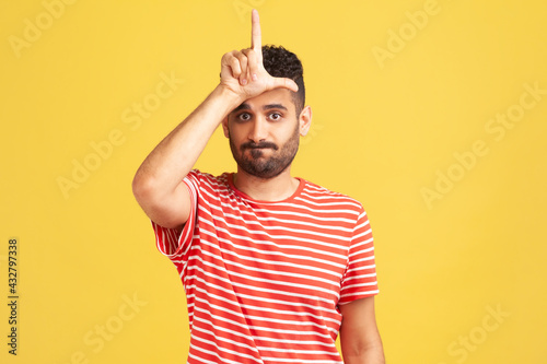 Gloomy depressed man with beard in striped t-shirt showing looser gesture holding fingers near forehead, sad because of silly mistake. Indoor studio shot isolated on yellow background photo