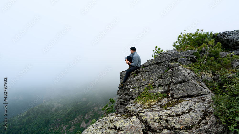 A man guy sits by a cliff on a rock in the fog in the mountains in summer