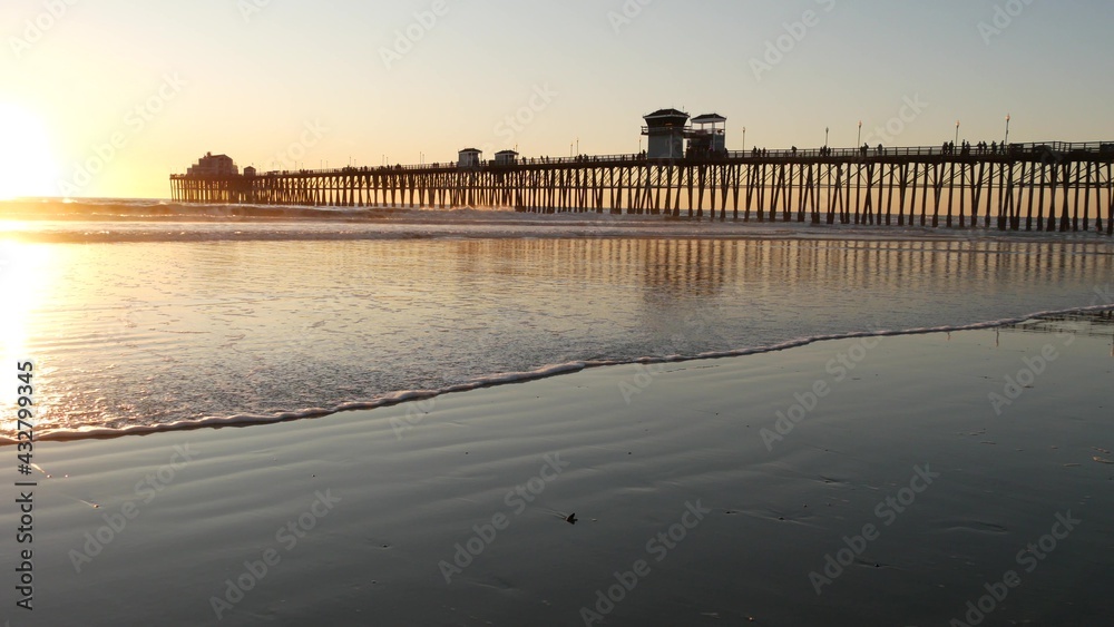 Wooden pier on piles, silhouette at sunset, California USA, Oceanside. Waterfront resort, pacific ocean tide, tropical beach. Summertime coastline vacations atmosphere. Sunny sea waves at sundown.