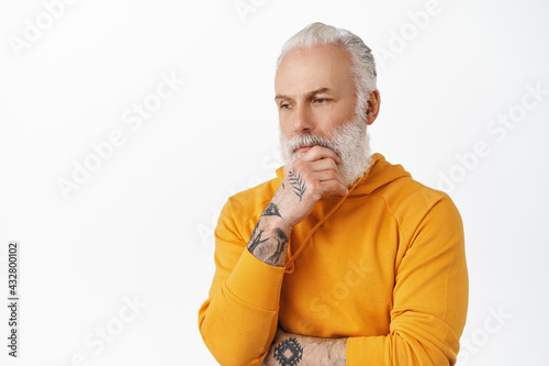 Stylish thoughtful mature man touch his beard, looking down and thinking serious, pondering important thing, standing pensive against white background