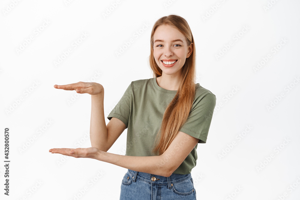 Young smiling female model showing an item, holding something, empty space between hands, looking pleased, recommending product, standing over white background Young smiling female model showing an item, holding something, empty space between hands, looking pleased, recommending product, standing over white background